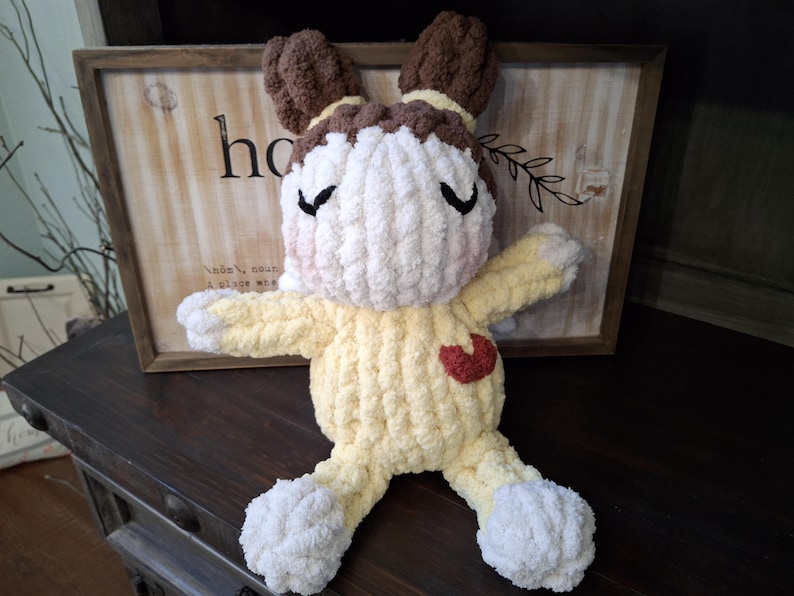 May include: A handmade, plush bunny doll with a yellow body, white head and feet, and brown ears. The doll has a red heart on its chest and closed black eyes. The background includes a wooden sign with the word "home".