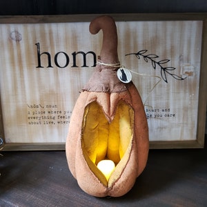 May include: A fabric pumpkin decoration in a muted orange hue with a brown stem and a heart-shaped cutout in the front. A small, glowing, battery-operated tea light sits inside the hollow center. A small circular tag with a black cat illustration is tied to the stem with twine.