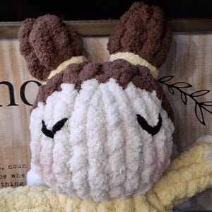 May include: A handmade, plush doll with a round face and a yellow body. The doll's hair is brown, and it has black eyes. The face is white with a touch of pink blush. The doll is made of soft, textured yarn, and is a decorative item.