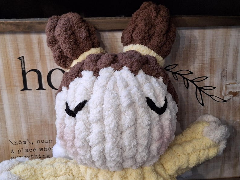 May include: A handmade, plush doll with a round face and a yellow body. The doll's hair is brown, and it has black eyes. The face is white with a touch of pink blush. The doll is made of soft, textured yarn, and is a decorative item.