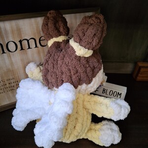 May include: A handmade crochet bunny in brown, white, and yellow. The bunny has floppy ears, a fluffy white tail, and yellow paws. The word "BLOOM" is visible on a small wooden block.