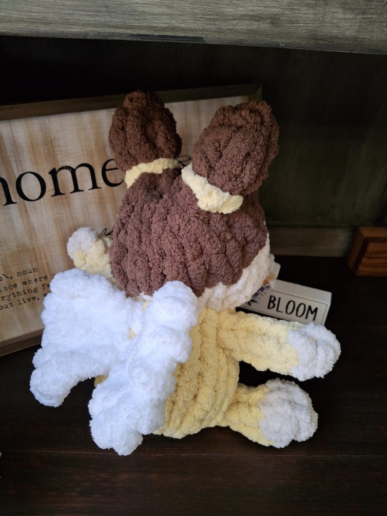 May include: A handmade crochet bunny in brown, white, and yellow. The bunny has floppy ears, a fluffy white tail, and yellow paws. The word "BLOOM" is visible on a small wooden block.