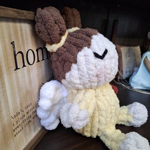May include: A handmade, plush bunny in shades of brown, white, and yellow. The bunny has floppy ears, a closed-eye design, and is sitting on a dark wooden surface. A wooden sign with the word "home" is in the background.