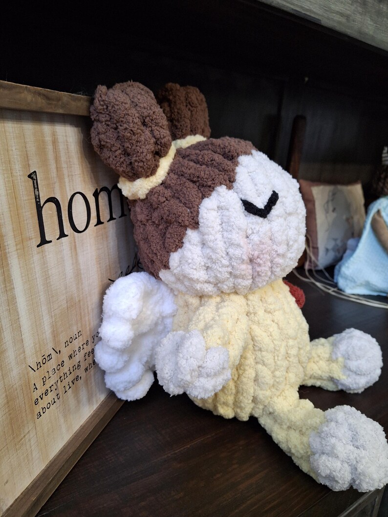 May include: A handmade, plush bunny in shades of brown, white, and yellow. The bunny has floppy ears, a closed-eye design, and is sitting on a dark wooden surface. A wooden sign with the word "home" is in the background.