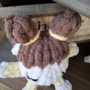 May include: A handmade, plush toy in the shape of a dog. The toy features brown, white, and yellow yarn. The dog has large, floppy ears and a fluffy body. The toy is resting on a wooden surface.