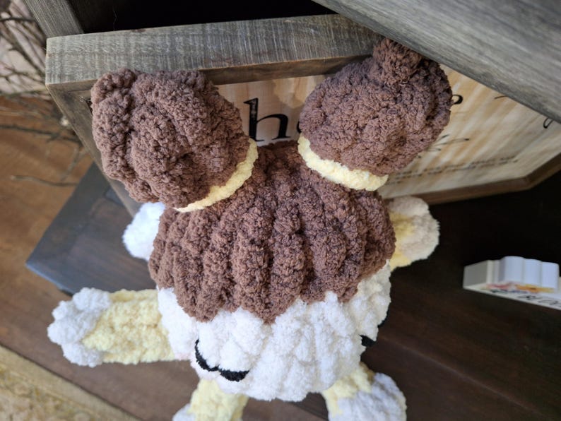 May include: A handmade, plush toy in the shape of a dog. The toy features brown, white, and yellow yarn. The dog has large, floppy ears and a fluffy body. The toy is resting on a wooden surface.