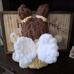 May include: A handmade, plush bunny toy with brown fur, yellow accents, and white fluffy ears and bow. The bunny is positioned in front of a wooden sign with the letter "h" visible. The toy is likely a decorative item or a child's toy.