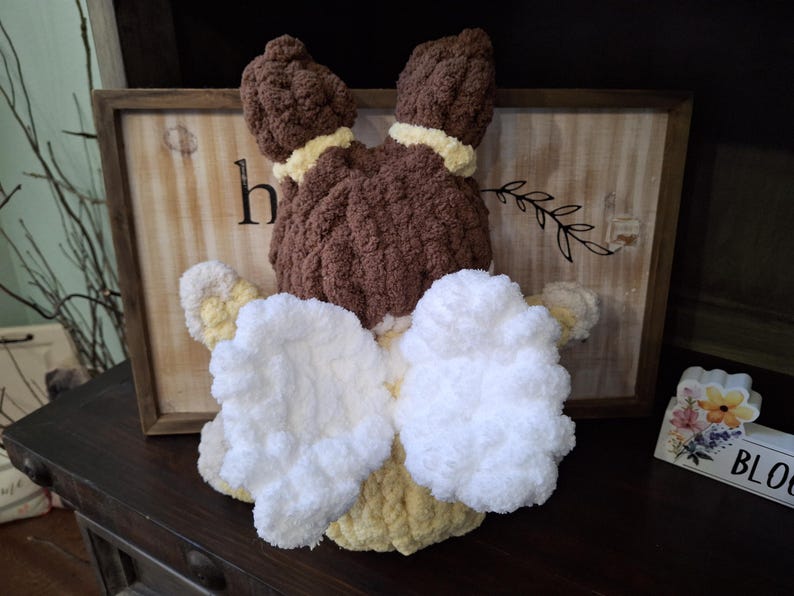 May include: A handmade, plush bunny toy with brown fur, yellow accents, and white fluffy ears and bow. The bunny is positioned in front of a wooden sign with the letter "h" visible. The toy is likely a decorative item or a child's toy.