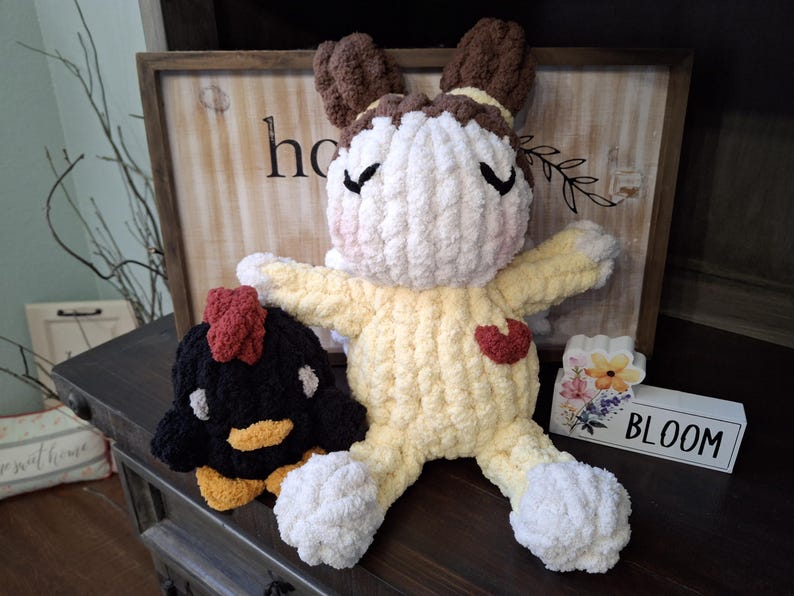 May include: A handmade, plush bunny doll with a yellow body, white limbs, and brown ears. A black and yellow chicken plush is next to the bunny. A wooden sign with the word "bloom" is also in the image.
