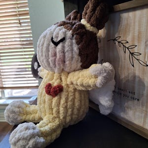 May include: A handmade crochet angel doll with a yellow body, white wings and feet, and a brown and white head. The doll has a red heart on its chest and a black stitched eye.