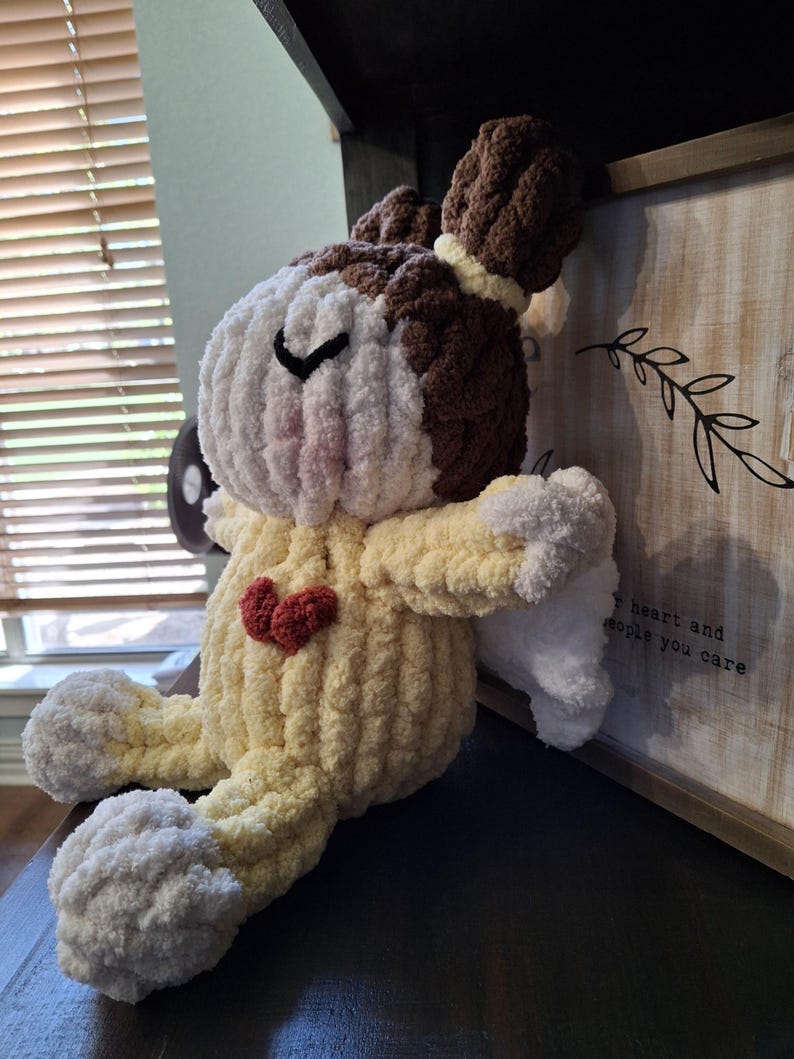 May include: A handmade crochet angel doll with a yellow body, white wings and feet, and a brown and white head. The doll has a red heart on its chest and a black stitched eye.