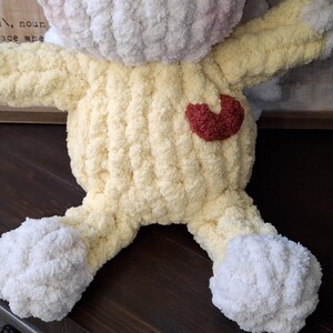 May include: A handmade, plush bunny in soft yellow, white, and pink yarn. The bunny has a red heart on its chest and is made with a chunky knit stitch. The toy is designed with arms and legs, perfect for a child's comfort.