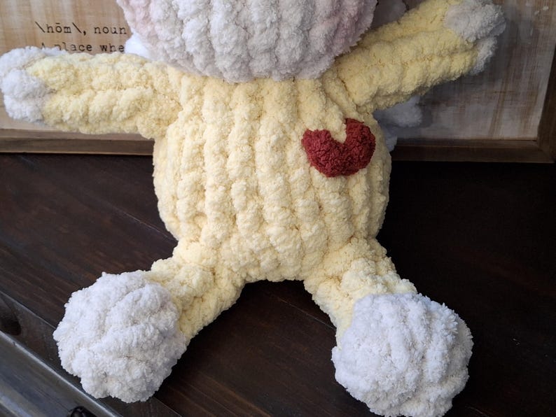 May include: A handmade, plush bunny in soft yellow, white, and pink yarn. The bunny has a red heart on its chest and is made with a chunky knit stitch. The toy is designed with arms and legs, perfect for a child's comfort.