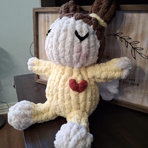 May include: A handmade, plush angel doll with a yellow body, white head and feet, and brown hair. The doll has a red heart on its chest and small white wings. The doll is sitting on a dark brown surface.