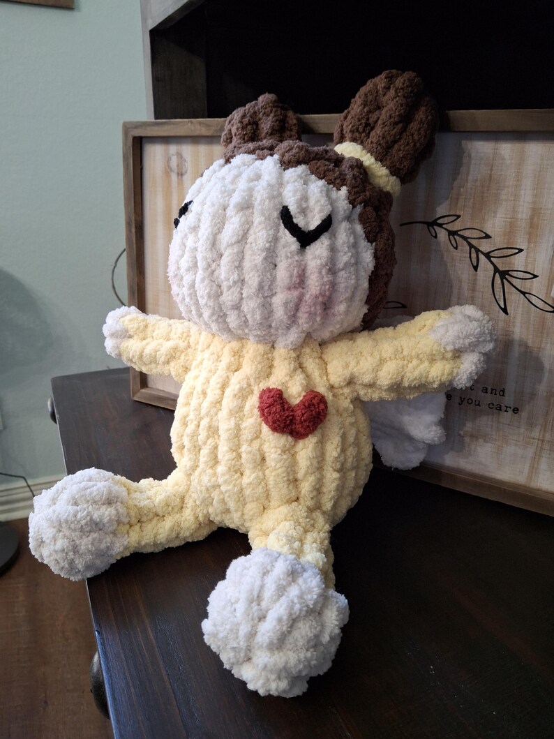 May include: A handmade, plush angel doll with a yellow body, white head and feet, and brown hair. The doll has a red heart on its chest and small white wings. The doll is sitting on a dark brown surface.