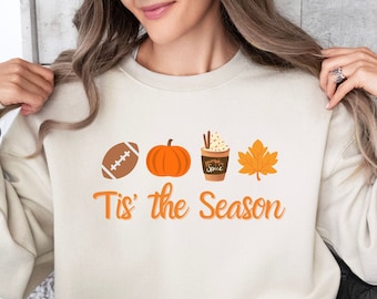 Puede incluir: Una sudadera beige con un diseño gráfico de un balón de fútbol americano, una calabaza, una taza de café con crema batida y una hoja de arce. El texto "Tis' the Season" está impreso debajo del gráfico.