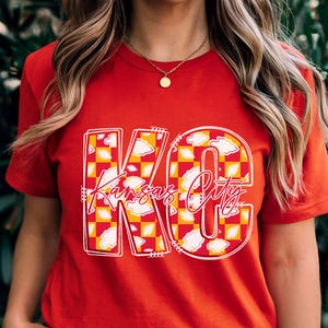 May include: Red t-shirt with a graphic design featuring the letters "KC" in white outline, filled with a yellow and red checkered pattern and white cloud accents. The words "Kansas City" are written in a script font.