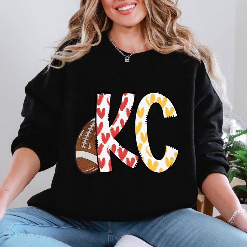 May include: Black sweatshirt featuring a football and the letters "KC" in a playful design. The "K" is decorated with red hearts, and the "C" is filled with yellow hearts. The football is brown and white.
