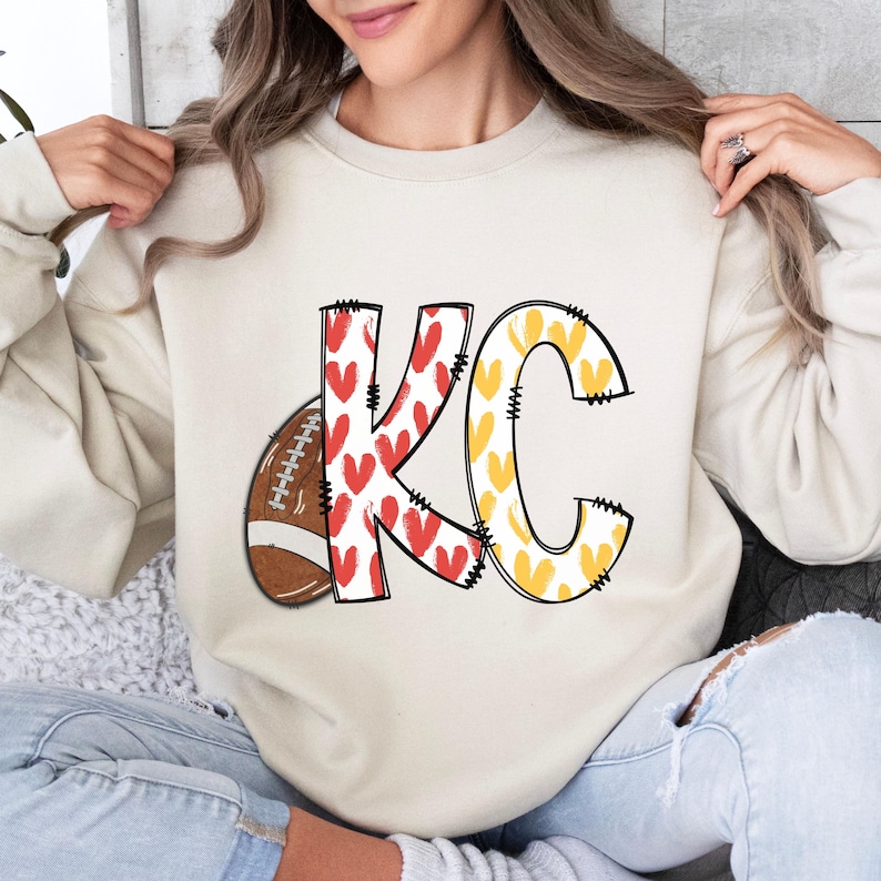 May include: Cream-colored sweatshirt featuring the letters "KC" in a playful design. The "K" is filled with red hearts, the "C" with yellow hearts, and a football graphic is on the left. The sweatshirt has a relaxed fit.