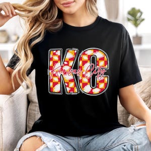 May include: Black t-shirt featuring the text "KC Kansas City" in a bold, decorative font. The letters are filled with a red and yellow checkered pattern and white cloud accents. The shirt is a casual style.