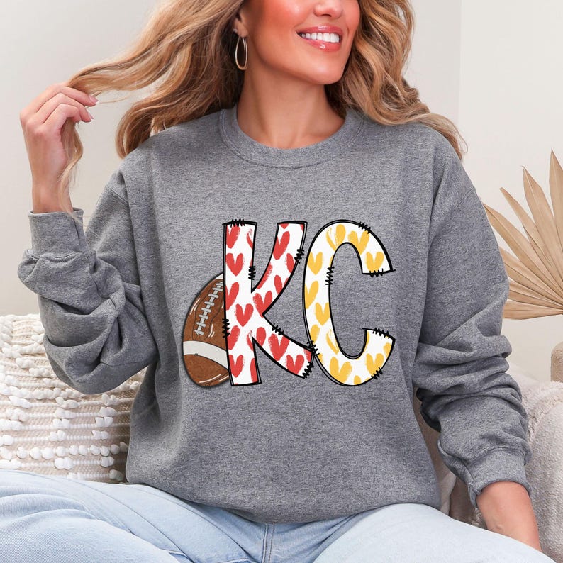 May include: Gray sweatshirt with the letters "KC" in a football-themed design. The "K" is decorated with red hearts, the "C" with yellow hearts, and a football is on the left side. The sweatshirt is a crew neck.