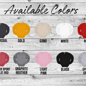 May include: A wooden background displays a variety of crewneck sweatshirts in various colors. The available colors are charcoal, gold, sand, white, red, heather sport scarlet red, graphite heather, light pink, black, and sport grey.
