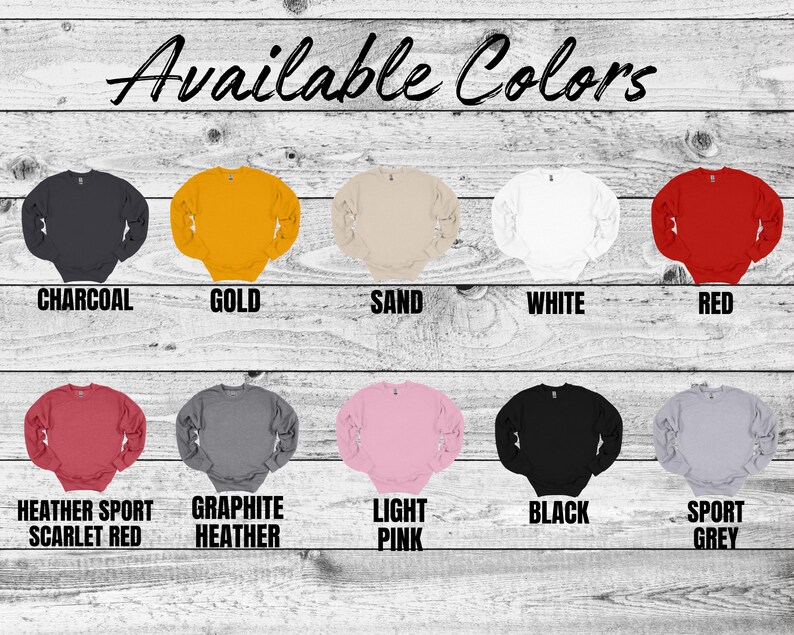 May include: A wooden background displays a variety of crewneck sweatshirts in various colors. The available colors are charcoal, gold, sand, white, red, heather sport scarlet red, graphite heather, light pink, black, and sport grey.