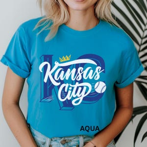 Puede incluir: Camiseta azul aguamarina con las palabras "Kansas City" en escritura blanca y un gr&aacute;fico de b&eacute;isbol. El dise&ntilde;o incluye una corona y las letras "KC" en azul. La palabra "AQUA" est&aacute; impresa en la parte inferior.