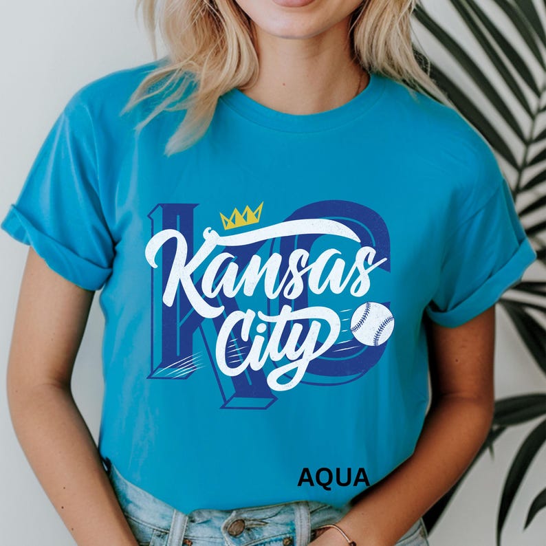 Puede incluir: Camiseta azul aguamarina con las palabras "Kansas City" en escritura blanca y un gr&aacute;fico de b&eacute;isbol. El dise&ntilde;o incluye una corona y las letras "KC" en azul. La palabra "AQUA" est&aacute; impresa en la parte inferior.