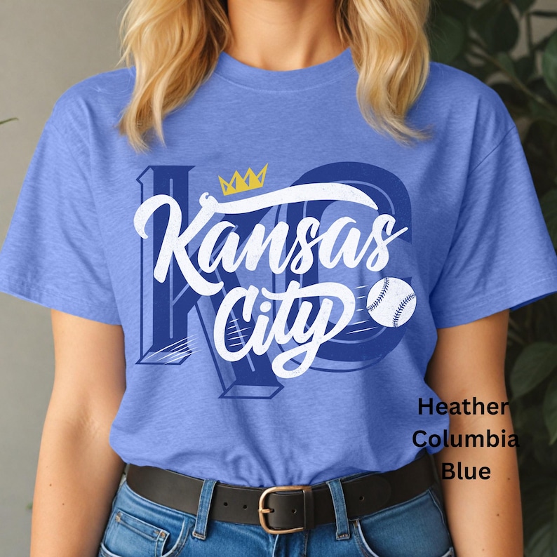 Puede incluir: Una camiseta azul con las palabras "Kansas City" en escritura blanca, una pelota de b&eacute;isbol y una corona dorada. La camiseta es de color azul Columbia jaspeado.
