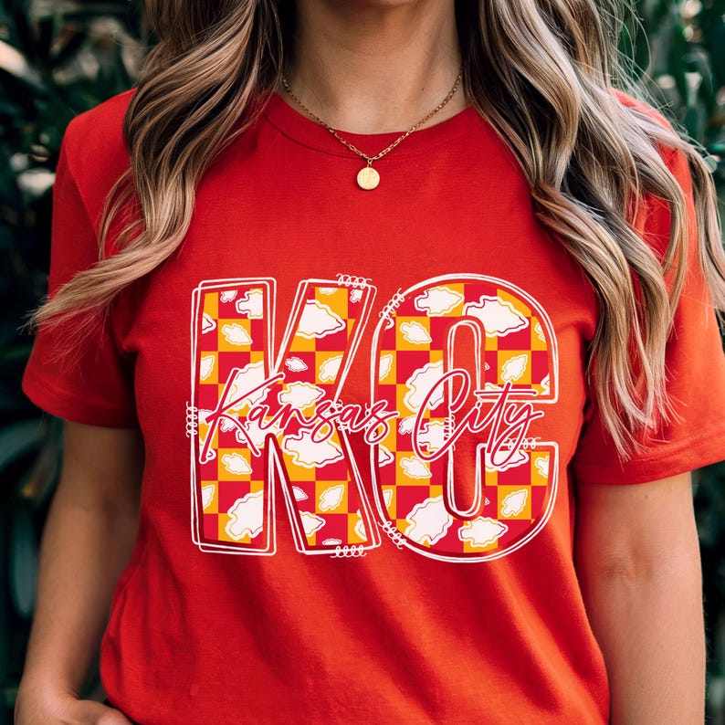May include: Red t-shirt with a graphic design featuring the letters "KC" in white outline, filled with a yellow and red checkered pattern and white cloud accents. The words "Kansas City" are written in a script font.