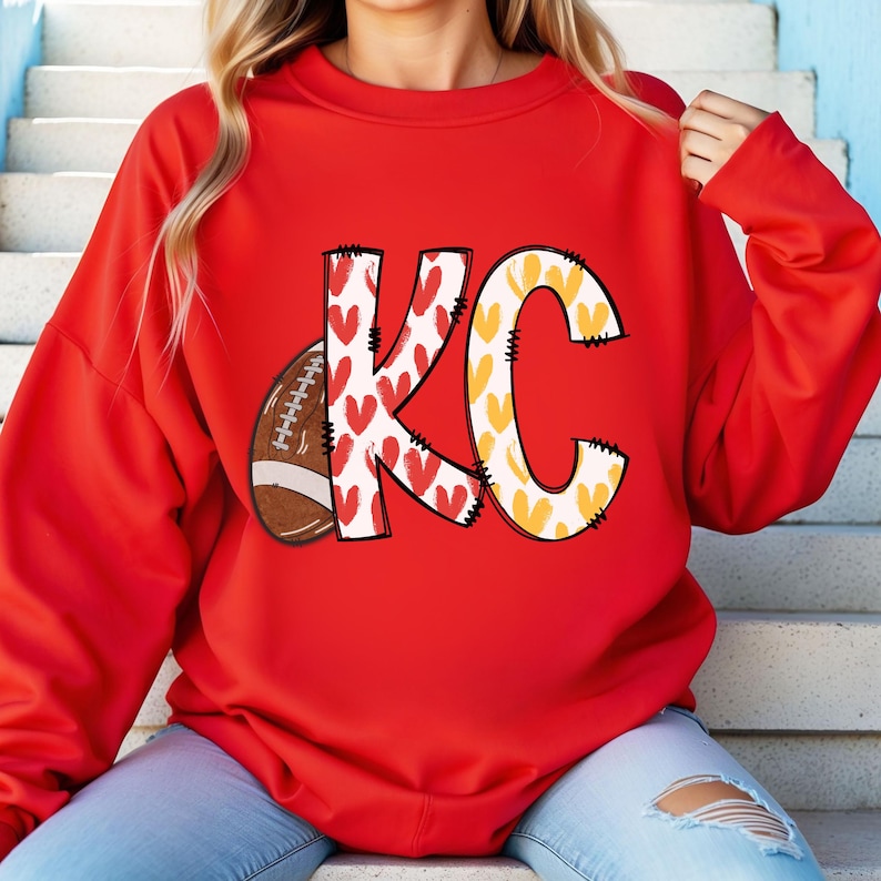 May include: A red sweatshirt featuring the letters "KC" in a playful design. The "K" is decorated with red hearts, while the "C" is adorned with yellow hearts. A football graphic is on the left side of the "K".