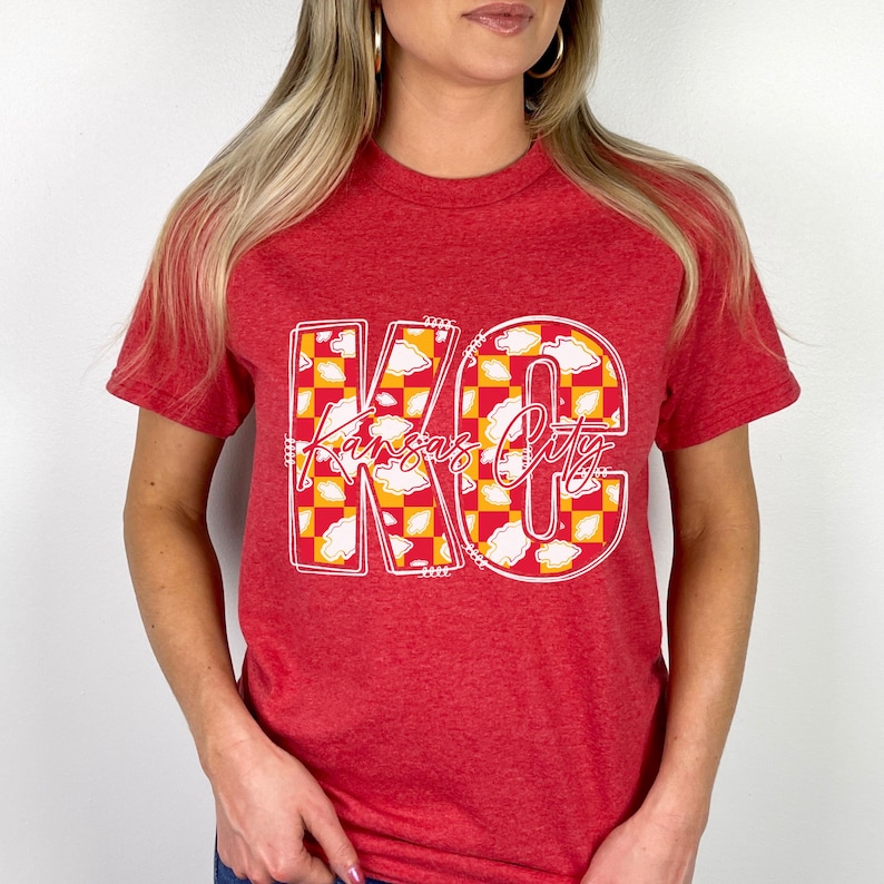 May include: Red t-shirt with the words "Kansas City" in a white outline. The letters are filled with a yellow and white checkered pattern and cloud designs.