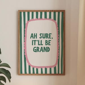 Könnte beinhalten: Ein gerahmter Druck mit dem Text "AH SURE, IT'LL BE GRAND" in grünen Buchstaben, vor einem weißen und rosa wellenförmigen Hintergrund. Der Rahmen hat grüne und weiße vertikale Streifen. Eine runde, leuchtende Lampe steht auf einem Holzregal.