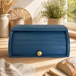 May include: A navy blue wooden bread bin with a gold knob. The bin has a roll-top design and is on a wooden surface. Sliced bread rests on a wooden board next to a linen cloth.