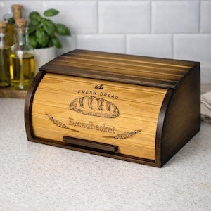 May include: Wooden bread bin with a curved lid and dark brown trim. The front of the bin is light brown and features the words "The Fresh Bread" and "Breadbasket" with a bread illustration.