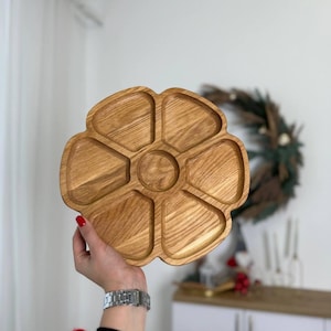 Puede incluir: Una bandeja de madera para servir con forma de flor. Está dividida en siete secciones, con un área central circular y seis compartimentos en forma de pétalo. La madera tiene una veta natural y un color marrón claro cálido.