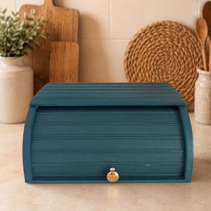 May include: A dark teal wooden bread box with a roll-top door and a small, round, gold-colored knob. The rectangular container features horizontal slats across the front panel and a smooth, flat top surface, providing a storage solution for bread.