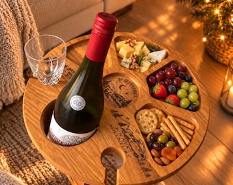 May include: A round wooden snack and wine table with a bottle holder, a glass holder, and three carved compartments filled with cheese, grapes, strawberries, crackers, breadsticks, olives, and nuts. The wood features engraved text and mountain art.