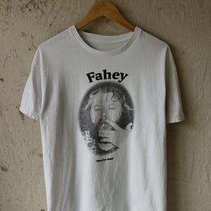 Op de afbeelding: Wit t-shirt met een zwart-wit afbeelding van een gezicht en een hand, met de naam "Fahey" boven de afbeelding. De tekst "Blind Joe Death" staat eronder. Het t-shirt hangt aan een houten hanger.