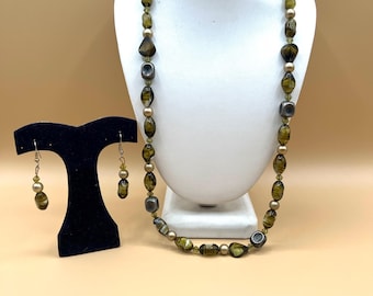 Könnte beinhalten: Ein Halsketten- und Ohrring-Set, das auf einer weißen Büste und einem schwarzen Ständer ausgestellt ist. Die Halskette hat olivgrüne und graue Perlen mit goldenen Akzenten. Die Ohrringe haben passende olivgrüne und goldene Perlen.