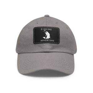 May include: A grey baseball cap featuring a rectangular black patch stitched onto the front. The patch displays a white graphic of a fish jumping out of water to catch a lure, with the text FISHING ADVENTURE printed in white capital letters.