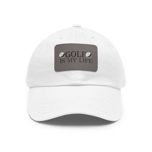 May include: A white baseball cap featuring a rectangular brown patch centered on the front. The patch displays the text GOLF IS MY LIFE in black lettering, flanked by two small illustrations of white golf balls on the left and right sides.