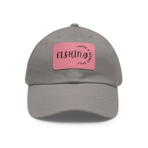 Puede incluir: Una gorra de béisbol gris con un parche rectangular rosa centrado en la parte delantera. El parche muestra el texto negro FISHING en una fuente cursiva grande, con las palabras I HAVE A SECRET SPOT curvadas en el lado derecho del parche.