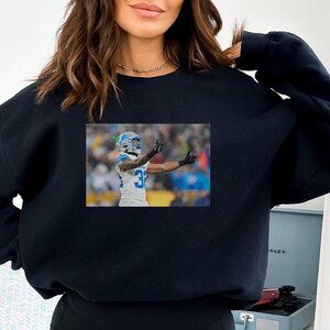 May include: A black sweatshirt with a graphic of a football player wearing a blue and white jersey, helmet, and white pants. The player is raising his right arm in the air.