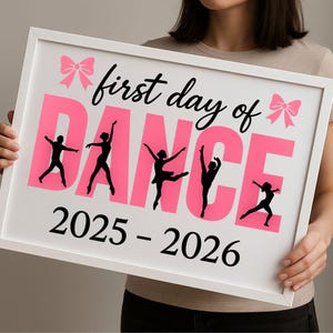 May include: A framed white sign with the text "first day of DANCE" in black script and pink block letters. Silhouettes of dancers are featured. The sign also includes the years "2025 - 2026" in black.