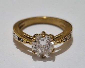 Op de afbeelding: Een gouden ring met een grote, heldere steen in het midden en kleinere, heldere stenen die in de band zijn gezet.