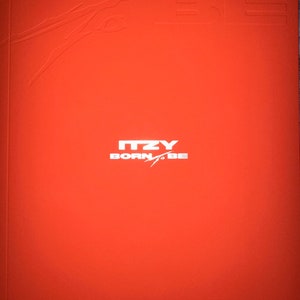 May include: A red album cover with the text "BORN TO BE" in white letters. The text is embossed and the album cover has a textured surface. The album cover also features the text "ITZY" in white letters.