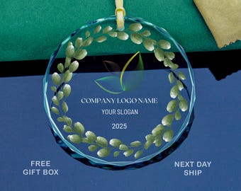 May include: A clear, circular glass ornament featuring a green leaf design and a company logo. The ornament is suspended by a yellow ribbon and displays the text "COMPANY LOGO NAME", "YOUR SLOGAN", and "2025".