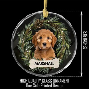 May include: A round, clear glass Christmas ornament with a festive green wreath design. A brown and white puppy is inside the wreath, with the name "MARSHALL" on a bone-shaped tag. The ornament is 3 1/5 inches in diameter.